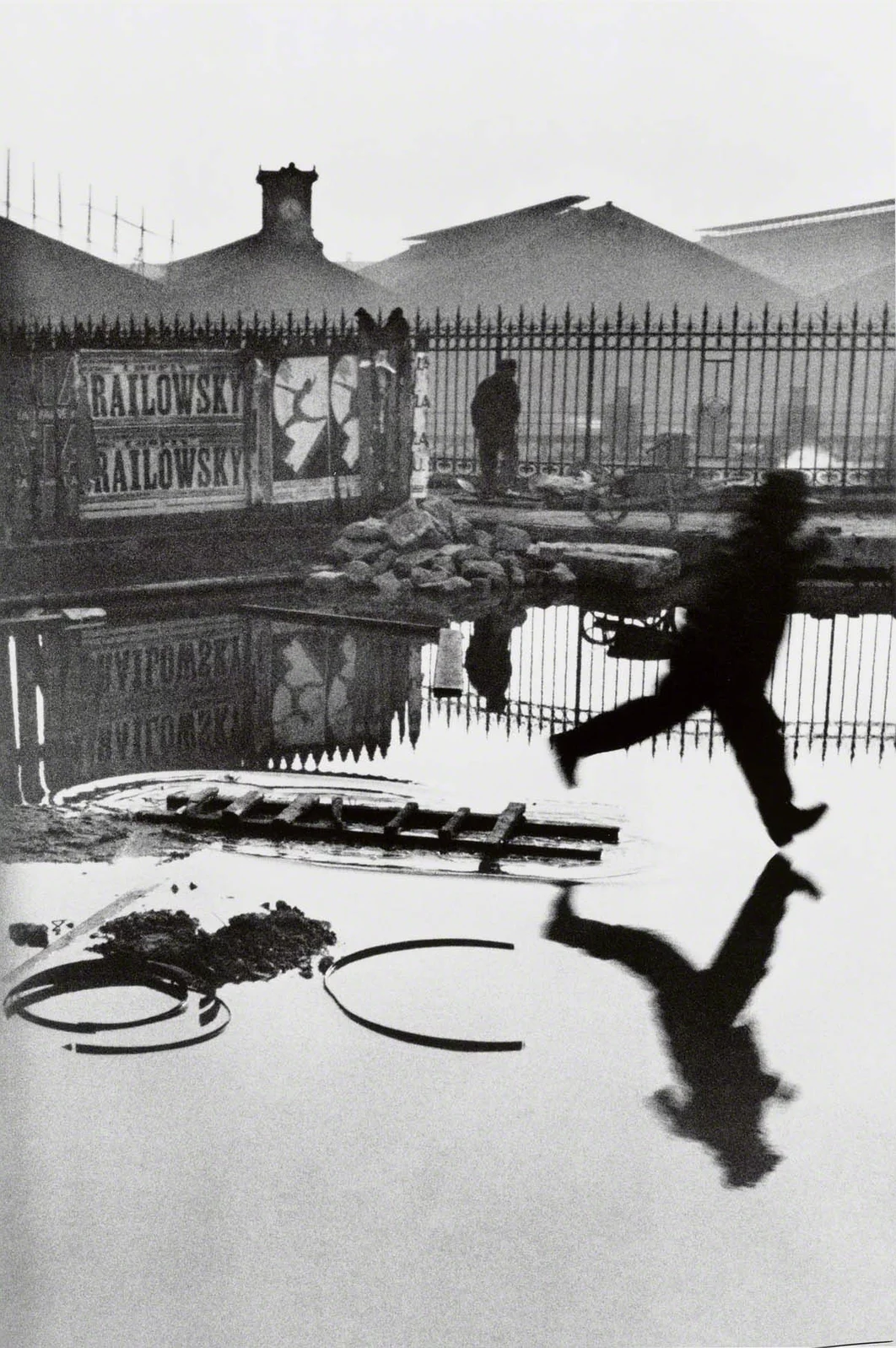 A man leaps over a flooded construction site behind the Gare Saint-Lazare, his body and reflection echoing a jumping figure on a poster beyond the fence.