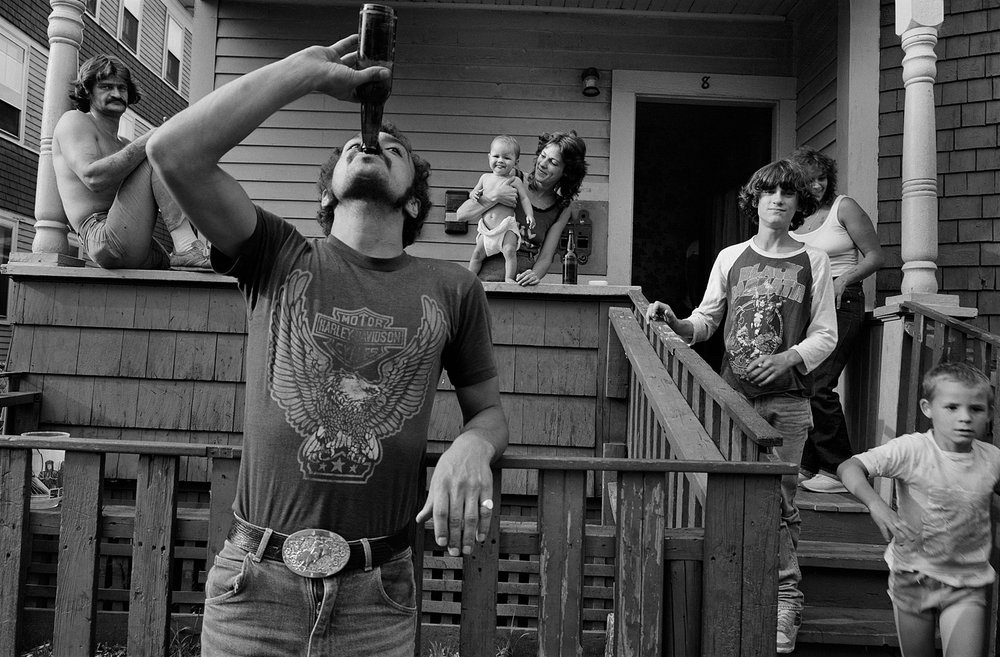 A man tilts back a bottle in front of a porch where adults and children gather across the steps, railings and doorway.