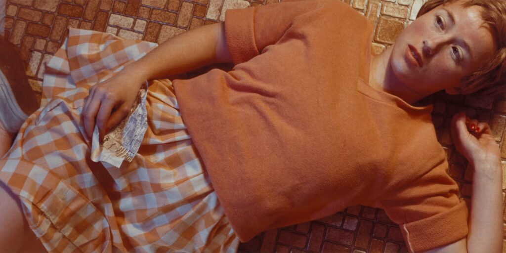 Cindy Sherman reclining on a harvest-gold floor in orange clothing and holding a newspaper clipping in Untitled #96.
