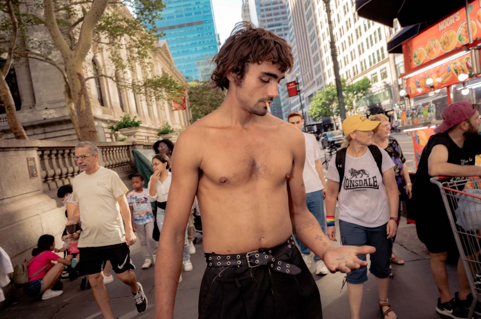 A shirtless man with a cigarette looks down at coins in his open palm on a crowded Midtown sidewalk beside a food cart.